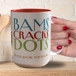 May include: White ceramic mug with a red interior, featuring the words "BAMS CRACKS DOTS" in teal, red, and green. The phrase "IF YOU KNOW, YOU KNOW" is printed below. The mug is on a wooden surface.