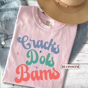 May include: A light pink t-shirt with the words "Cracks Dots Bams" in blue, green, and coral lettering. The shirt has a crew neck and a tag that says "Comfort Colors". A tan hat and blue jeans are in the background.