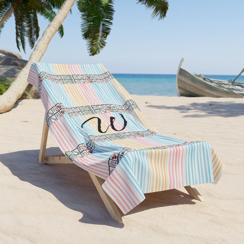 May include: A beach towel featuring a pastel striped pattern in light blue, yellow, and pink. The towel displays a horizontal band of mahjong tiles and a large, black, cursive letter W centered on the fabric, draped over a wooden lounge chair.