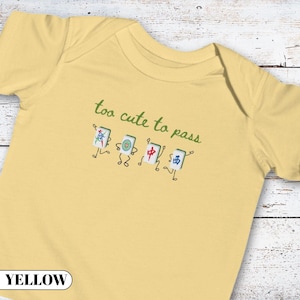 May include: A pale yellow short-sleeved baby bodysuit featuring a graphic of four animated mahjong tiles with arms and legs. Above the tiles, the text reads "too cute to pass" in green cursive lettering. The tiles display various symbols.