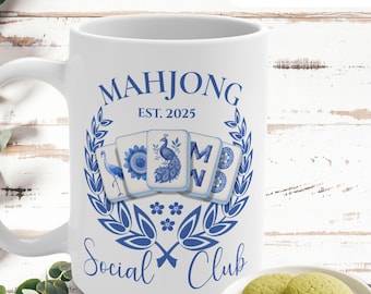 May include: A white ceramic mug featuring a blue graphic design with the text "MAHJONG EST. 2025 SOCIAL CLUB" surrounding five mahjong tiles decorated with blue illustrations of a crane, a flower, a peacock, the letter M, and a circular pattern.