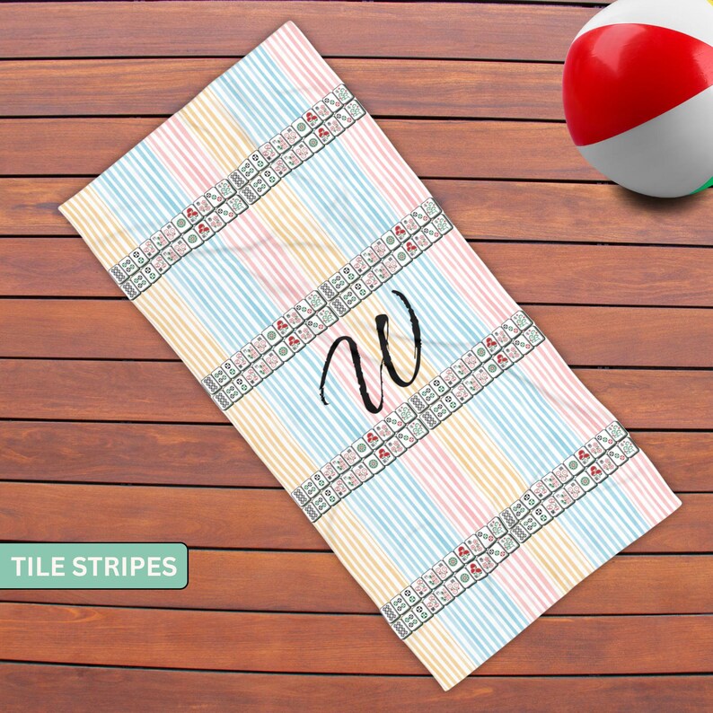 May include: A rectangular beach towel featuring vertical stripes in pastel pink, blue, and yellow. Two horizontal bands of mahjong tile patterns cross the towel, and a large, black, handwritten-style letter W is centered between the tile bands.