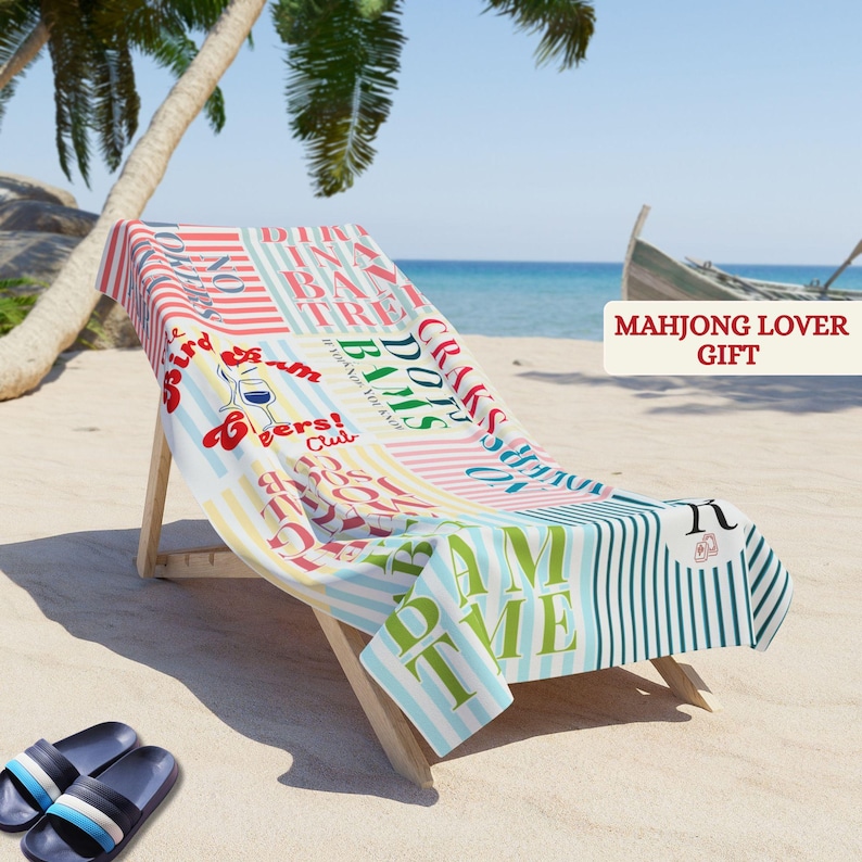 May include: A beach towel draped over a wooden lounge chair, featuring a multi-colored striped pattern with various printed phrases including "BAMS," "DOTS," "CRACKS," and "MAHJONG LOVER GIFT." The towel has sections of red, blue, and green text.