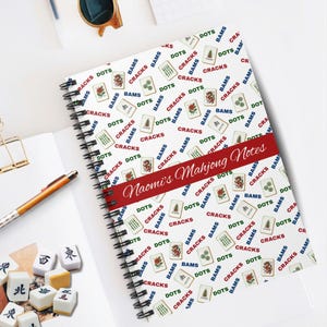 May include: Spiral-bound notebook with a white cover featuring a pattern of Mahjong tiles in red, green, and blue. The notebook has a red band with the text "Naomi's Mahjong Notes". A pen, sunglasses, and Mahjong tiles are also visible.