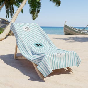 May include: A beach towel featuring a light blue and white vertical striped pattern. The towel displays a circular emblem with a black letter R in the center, flanked by two smaller circular icons showing a deck of cards and a coffee mug.