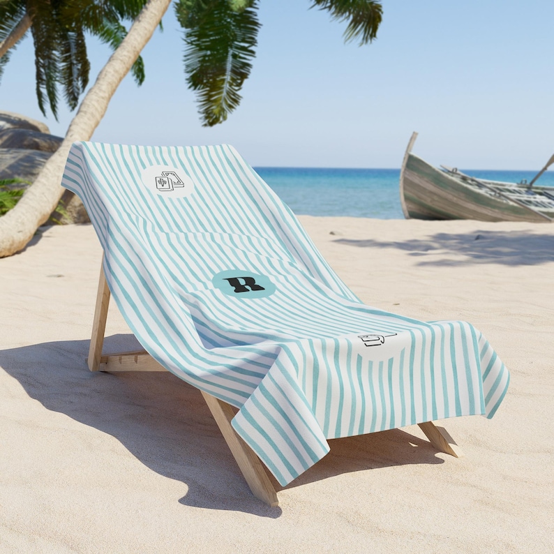 May include: A beach towel featuring a light blue and white vertical striped pattern. The towel displays a circular emblem with a black letter R in the center, flanked by two smaller circular icons showing a deck of cards and a coffee mug.