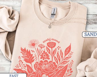 May include: A sand-colored crewneck sweatshirt featuring a coral-colored graphic of a floral bouquet. The design incorporates the text "mahj mood" and various Mahjong-related terms such as "bird," "bam," "cheers," "suits," "honors," "craks," "dots," "winds," "dragons," "jokers," "flowers," "kong," "pung," "chow," "quint," and "a joker" integrated into the leaves and petals.
