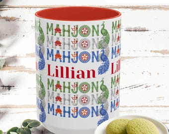 May include: A white ceramic mug with a red interior, featuring the name "Lillian" in red. The mug is decorated with the word "MAHJONG" in various colors, peacock illustrations, and the text "LOVELY GIFT". Green cookies are on a white plate.