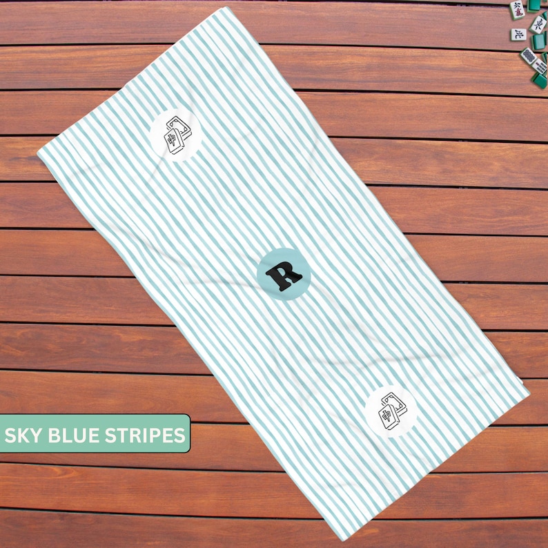 May include: A rectangular towel featuring a pattern of thin sky blue stripes on a white background. The towel includes a central circular emblem with the letter R and two smaller circular icons displaying a pair of mahjong tiles in black ink.