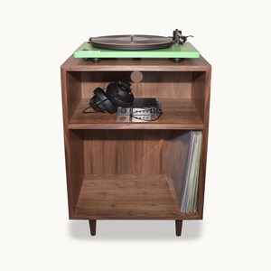 May include: A mid-century modern record player stand with a mint green turntable on top. The stand is made of dark wood and has two shelves. Black headphones and a small amplifier are on the middle shelf, and vinyl records are stored below.