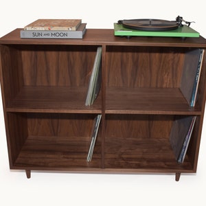 May include: A dark brown wooden record storage cabinet with four compartments. A green turntable sits on top, along with two books. Vinyl records are stored in the compartments. The cabinet has a mid-century modern design.