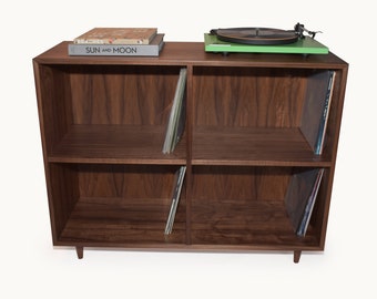 May include: A dark brown wooden record storage cabinet with four compartments. A green turntable sits on top, along with two books. Vinyl records are stored in the compartments. The cabinet has a mid-century modern design.