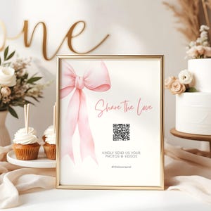 May include: A framed sign with a gold border features a pink bow illustration and the text "Share the Love." Below the text is a QR code and the words "Kindly send us your photos & videos." The sign is part of a party or event setup.