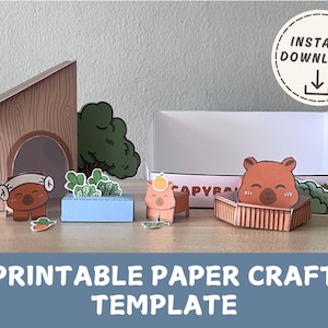 Peut inclure: Un modèle de bricolage en papier imprimable sur le thème du capybara. La scène comprend une maison, un arbre et une boîte, tous en papier. L'image comprend également un personnage de capybara et les mots "Téléchargement instantané".