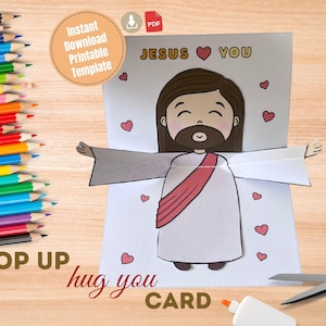 May include: A pop-up card featuring a cartoon illustration of Jesus with outstretched arms, surrounded by hearts. The card reads "JESUS YOU" and "hug you CARD". Colored pencils, scissors, and glue are also visible.