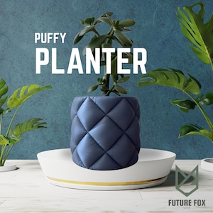 May include: A blue, quilted-effect planter containing a green plant, set on a white tray with a gold trim. The words "PUFFY PLANTER" are displayed in white. The planter has a diamond pattern.