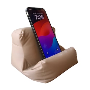 Puede incluir: Un soporte de teléfono de tela beige que sostiene un teléfono inteligente. El teléfono muestra una pantalla colorida con la hora 7:08. El soporte está diseñado para sostener un teléfono para una visualización con manos libres. El soporte está hecho de un material suave y acolchado.