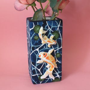 May include: A rectangular vase with a dark blue background featuring a white water ripple pattern, small green lily pads, and two raised, three-dimensional orange and white koi fish figures. The vase contains several pink artificial flowers.