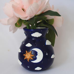 May include: A dark blue ceramic vase with a celestial design, featuring a sun, moon, stars, and clouds. The vase holds pink flowers with green leaves. The vase is approximately 15 cm tall.