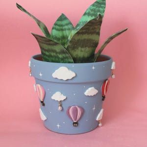 May include: A light blue plant pot featuring raised white clouds and colourful hot air balloons in pink, purple, and red. Small dangling pearl-like beads hang from the rim, and white star accents are painted across the surface of the pot.