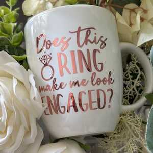 May include: White ceramic mug with a rose gold design that says "Does this ring make me look engaged?" The mug is surrounded by white flowers.