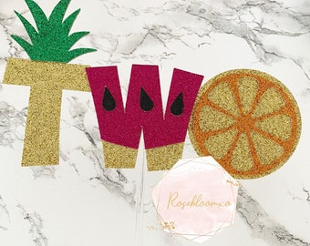 May include: A glittery cake topper with the word "TWO" spelled out with fruit-themed letters. The "T" is gold with a green pineapple top, the "W" is pink with black seed details, and the "O" is a gold orange slice.