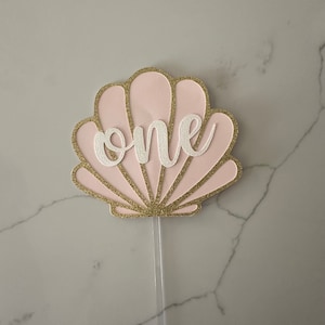 May include: Pink and gold glitter seashell cake topper with the word "one" in white lettering.