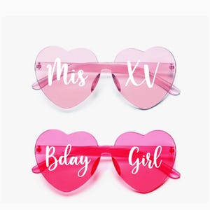 May include: Two pairs of heart-shaped sunglasses. One pair is light pink with "Mis XV" in white text. The other pair is a darker pink with "Bday Girl" in white text. Both pairs have clear frames.