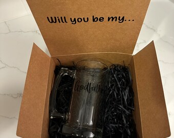 May include: A clear glass beer mug with the word "Godfather" printed on it, inside a brown cardboard box filled with black shredded paper. The box lid reads "Will you be my..." in black text.