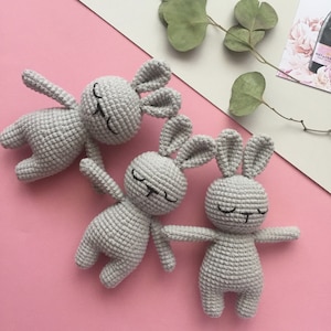 May include: Three gray crocheted bunny toys with black embroidered eyes and mouths. The bunnies are lying on a pink surface.