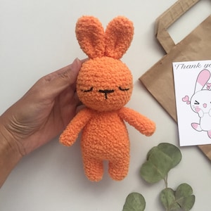 May include: A small orange crocheted bunny with closed eyes and a black embroidered nose. The bunny is sitting on a white surface.