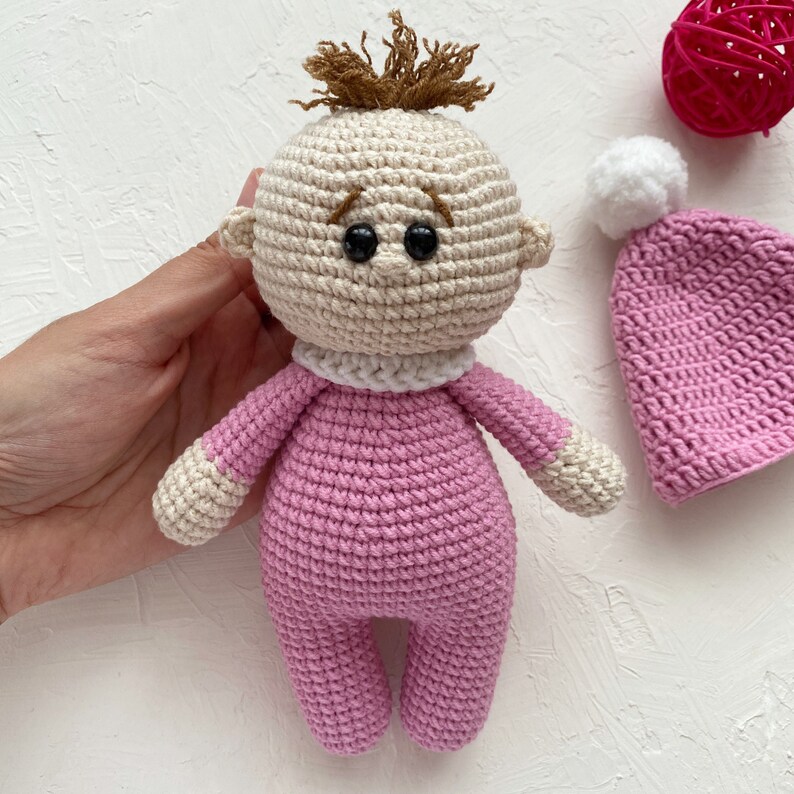 May include: A crocheted pink baby doll with brown yarn hair, black eyes, and a white crocheted collar. The doll is wearing a pink crocheted outfit and is holding a pink crocheted hat with a white pom-pom.