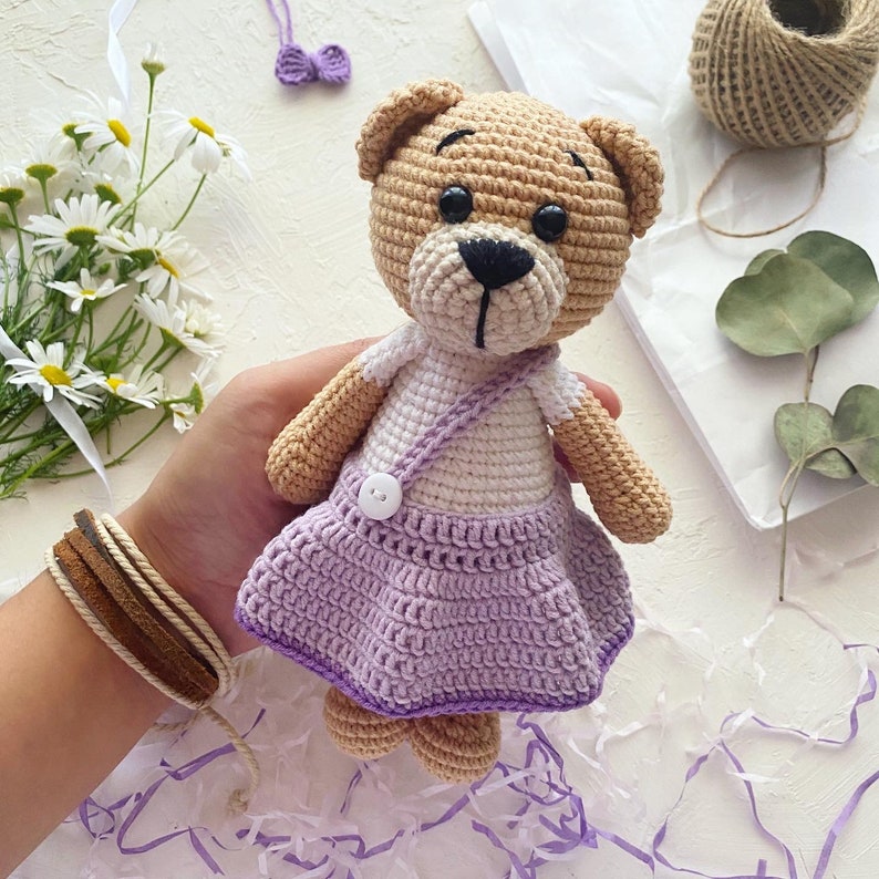 May include: A crocheted teddy bear wearing a purple and white dress. The bear has brown eyes and a black nose.