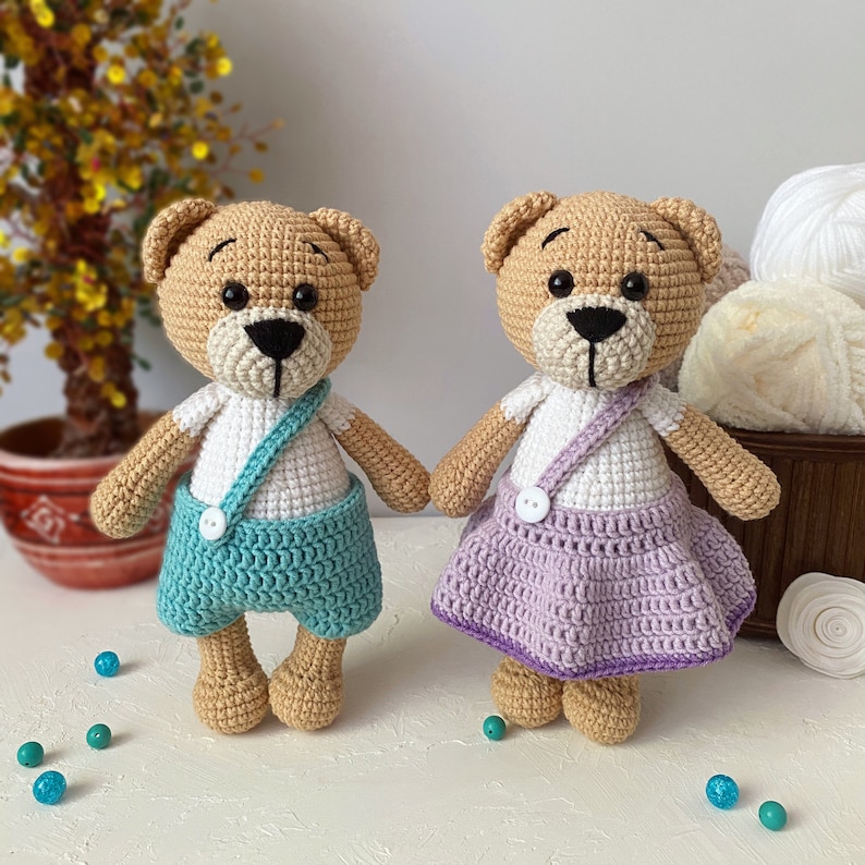 May include: Two crocheted teddy bears, one wearing a teal blue overall and the other wearing a lavender skirt. Both bears have white shirts and black eyes.
