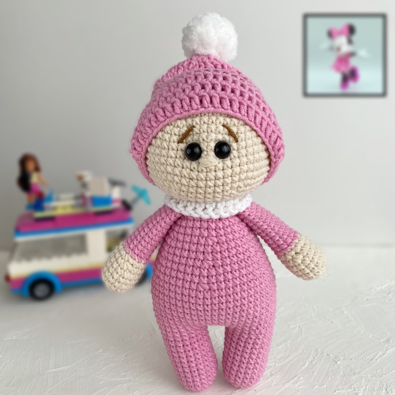 K&ouml;nnte beinhalten: Eine rosa geh&auml;kelte Puppe, die einen rosa und wei&szlig;en geh&auml;kelten Hut mit einem wei&szlig;en Bommel tr&auml;gt. Die Puppe hat braune Augen und einen wei&szlig;en geh&auml;kelten Kragen.