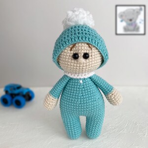 K&ouml;nnte beinhalten: Eine geh&auml;kelte Baby-Puppe mit einem blauen und wei&szlig;en Hut und einem wei&szlig;en Bommel. Die Puppe tr&auml;gt ein blaues H&auml;kel-Outfit.