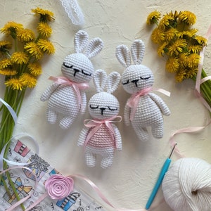 May include: Three white crocheted bunny rabbits with pink ribbon accents. The bunny in the center is wearing a pink crocheted dress. The bunnies are on a white surface with a bouquet of yellow flowers and a ball of white yarn.