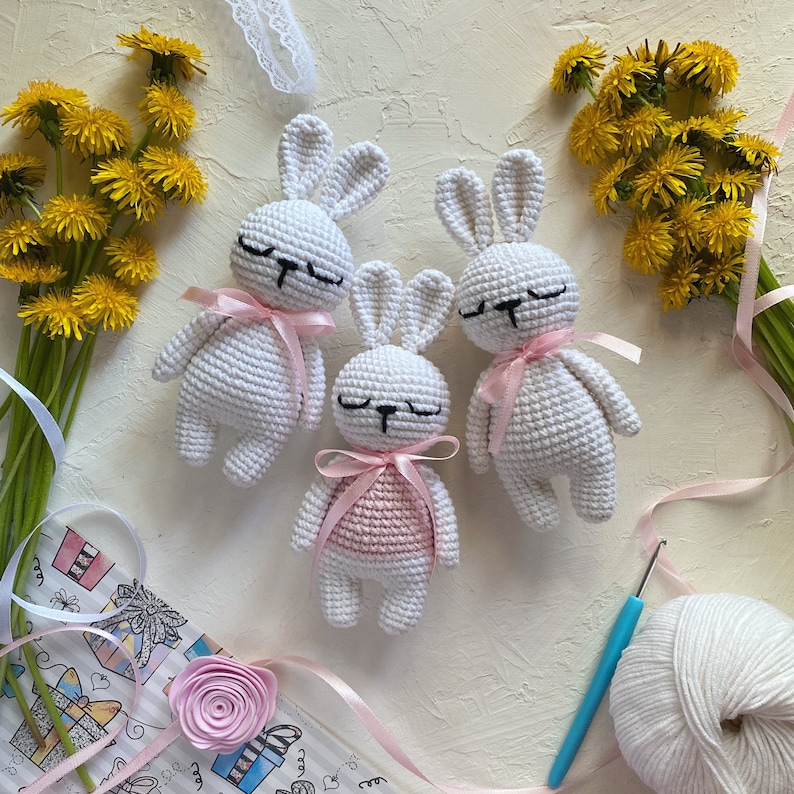 May include: Three white crocheted bunny rabbits with pink ribbon accents. The bunny in the center is wearing a pink crocheted dress. The bunnies are on a white surface with a bouquet of yellow flowers and a ball of white yarn.