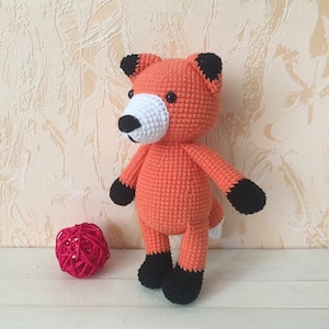May include: A crocheted orange fox plush toy with black ears, black feet, and a white nose. The toy is standing on a white surface.