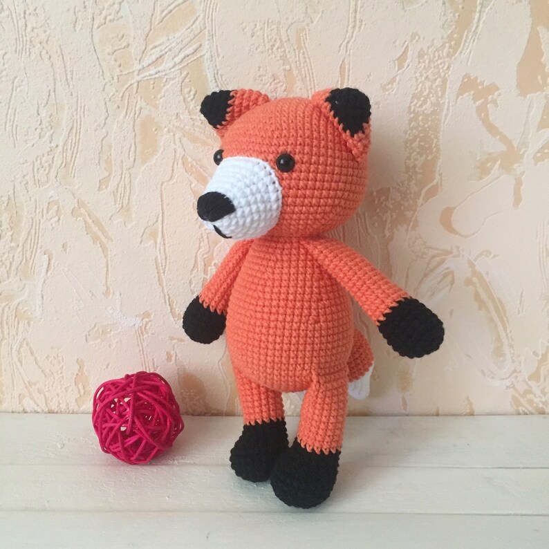 May include: A crocheted orange fox plush toy with black ears, black feet, and a white nose. The toy is standing on a white surface.