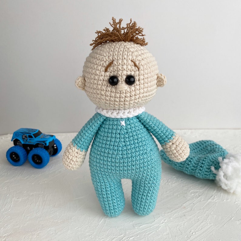 May include: A crocheted baby doll with brown hair, blue eyes, and a light blue outfit. The doll is wearing a white crocheted collar and has a matching light blue crocheted hat.