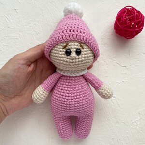 K&ouml;nnte beinhalten: Eine rosa geh&auml;kelte Babypuppe mit einem wei&szlig;en Bommelhut und braunen Augen.