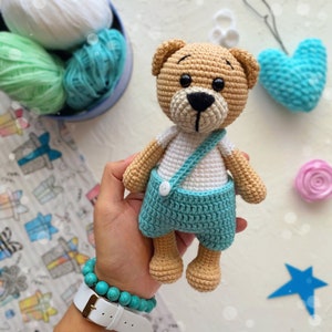 May include: A crocheted teddy bear wearing blue overalls with a white shirt and a single button. The bear has brown fur and black eyes.