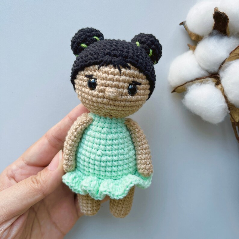 Puede incluir: Una mu&ntilde;eca de crochet con cabello negro, vestido verde y piel marr&oacute;n. La mu&ntilde;eca tiene una expresi&oacute;n dulce y lleva un vestido verde con un dobladillo con volantes.