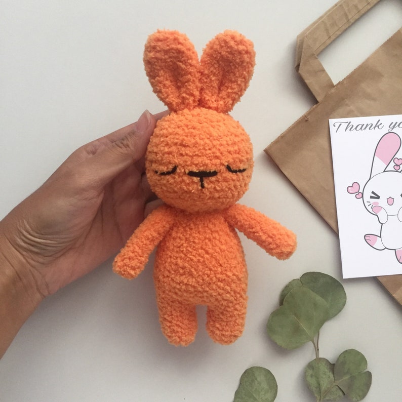 May include: A small orange crocheted bunny with closed eyes and a black embroidered nose. The bunny is sitting on a white surface.