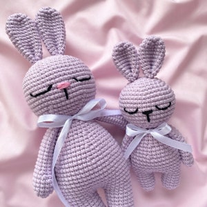 Puede incluir: Dos peluches de conejo de crochet de color lavanda con ojos negros y una nariz rosa. El conejo más grande tiene una cinta blanca atada alrededor de su cuello. El conejo más pequeño tiene una cinta blanca atada alrededor de su cintura.