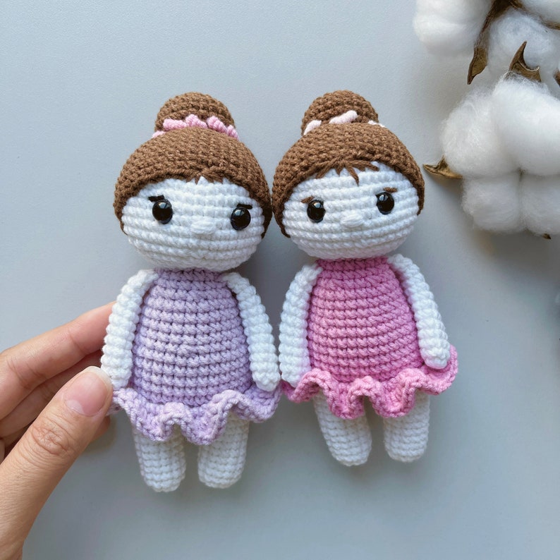 Puede incluir: Dos mu&ntilde;ecas de crochet con cabello marr&oacute;n y caras blancas que llevan vestidos rosa y morado. Las mu&ntilde;ecas tienen ojos negros y est&aacute;n de pie sobre una superficie blanca.