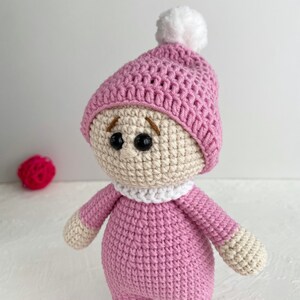 May include: A crocheted doll wearing a pink hat with a white pom-pom. The doll has a white crocheted collar and brown embroidered eyes.