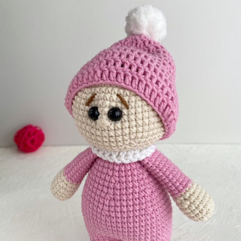 May include: A crocheted doll wearing a pink hat with a white pom-pom. The doll has a white crocheted collar and brown embroidered eyes.