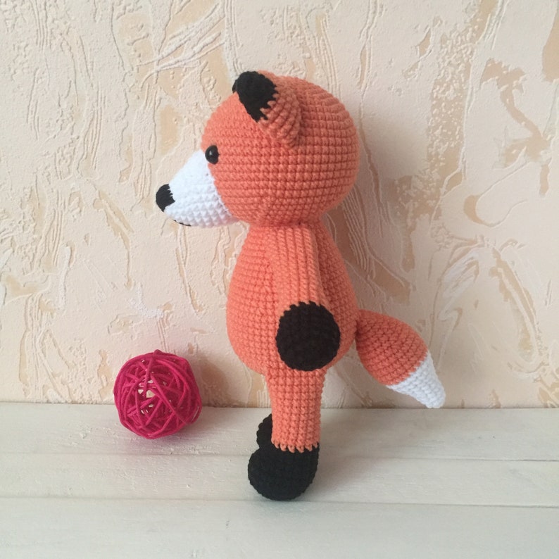 May include: A crocheted orange fox toy with black ears, nose, and feet. The fox has a white tail tip and a black spot on its leg.